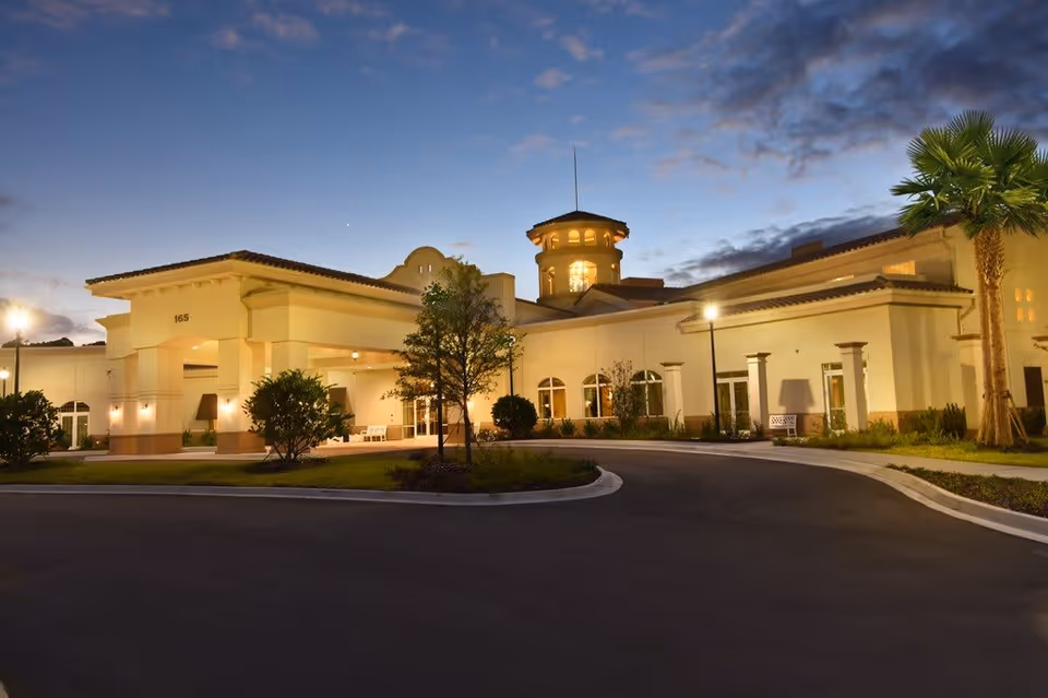 Exterior view of the Silver Creek senior living facility at dusk, showing a well-lit building with a covered entrance, arched windows, a tower feature, palm trees, and a curved driveway.