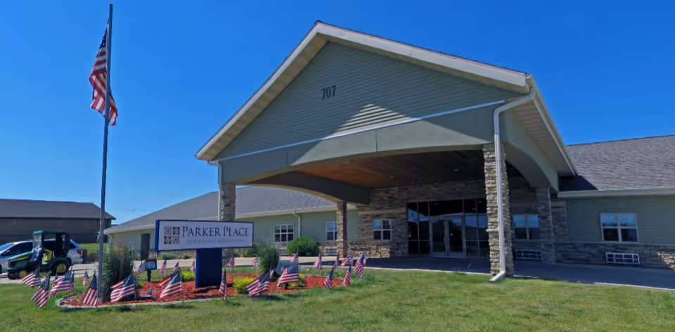 Front entrance of Parker Place Retirement Community with a covered portico, a sign on the lawn and multiple American flags.