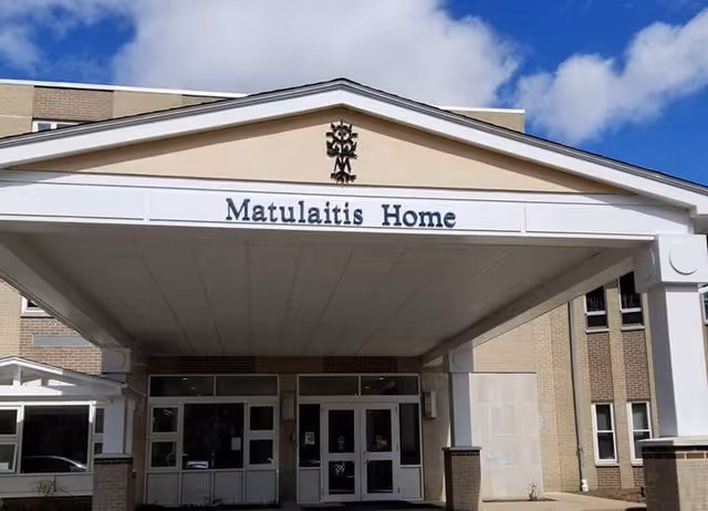 Entrance of Matulaitis Home nursing facility with a covered drop-off area, beige brick exterior, and a clear blue sky with some clouds above.
