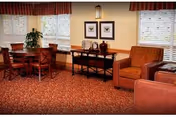 A cozy interior room with a red patterned carpet, two brown armchairs, a wooden table with four chairs, a black console table with decorative items, two framed butterfly pictures on the wall, and windows with white blinds and red valances.