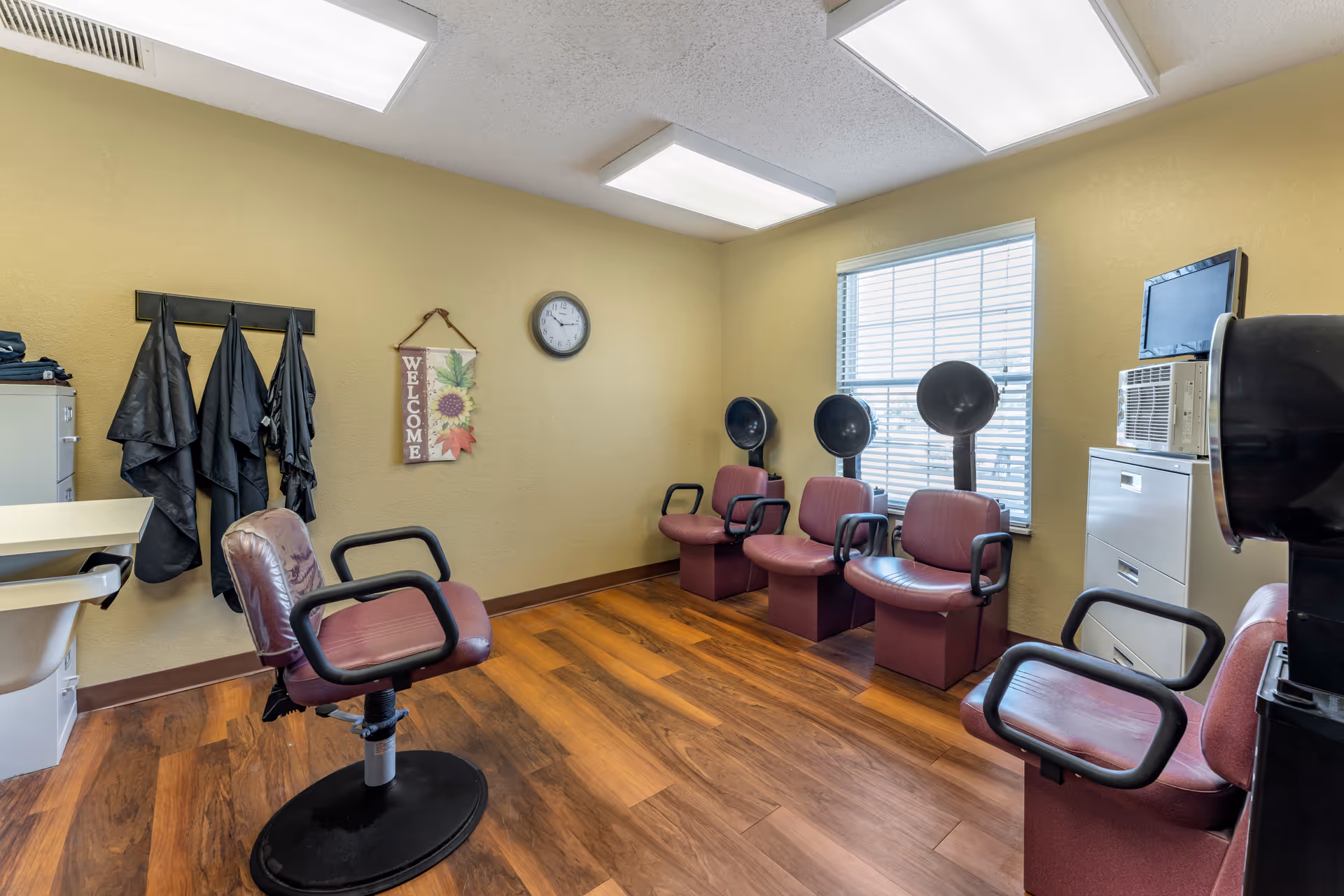 Interior view of a small salon room with four maroon salon chairs, three with hair dryers attached, and one standalone chair. The room has wood flooring, yellow walls, a window with blinds, a clock on the wall, a welcome sign, and black salon capes hanging on hooks.