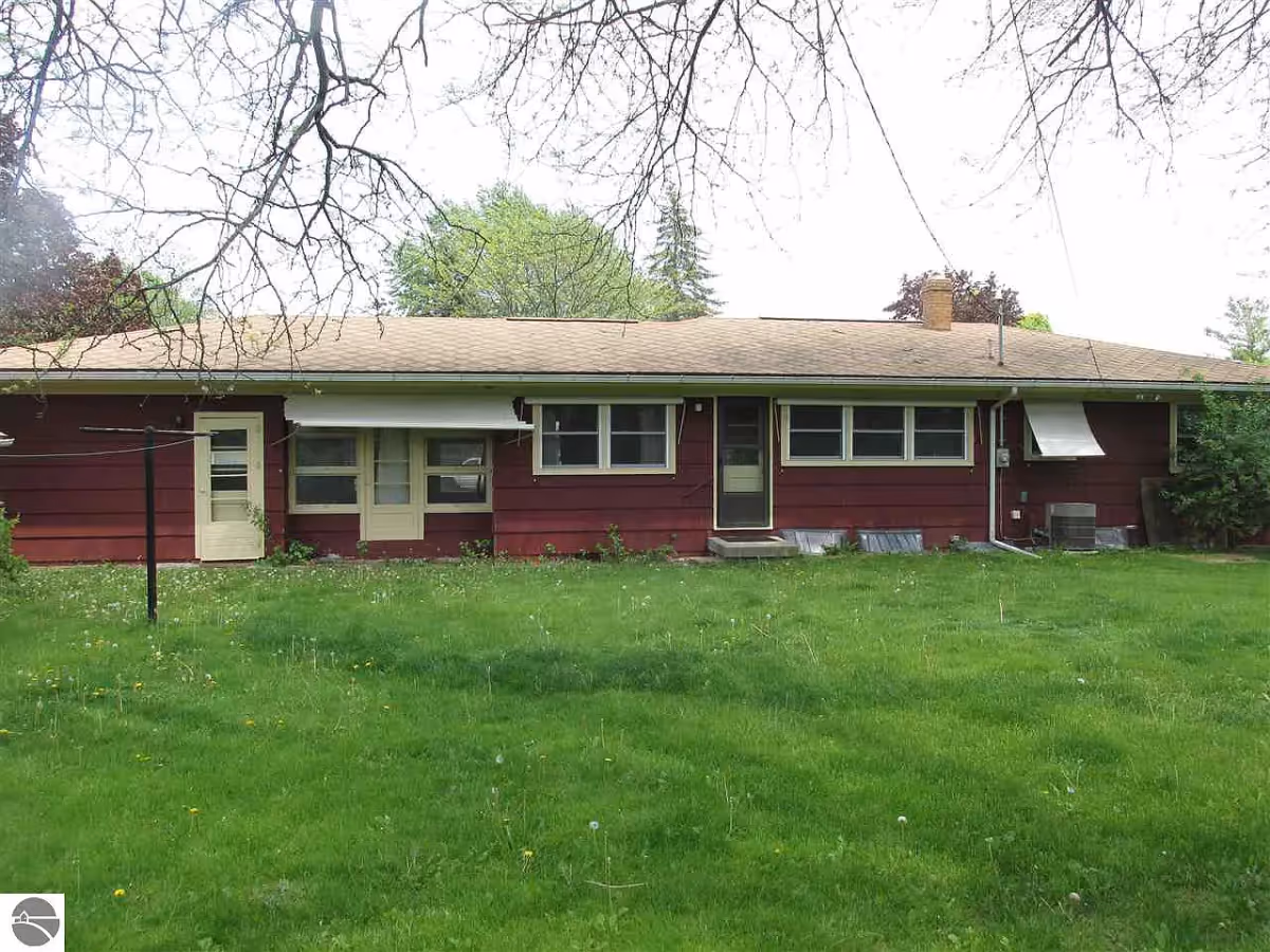Rear exterior view of a single-story red house with a beige roof, multiple windows, and two doors opening to a green grassy yard. Leafless tree branches are visible at the top of the image.