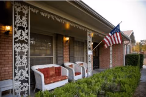 Front porch of a brick building with white decorative columns, two white wicker chairs with red cushions, an American flag mounted on a pole, and green shrubbery in front.