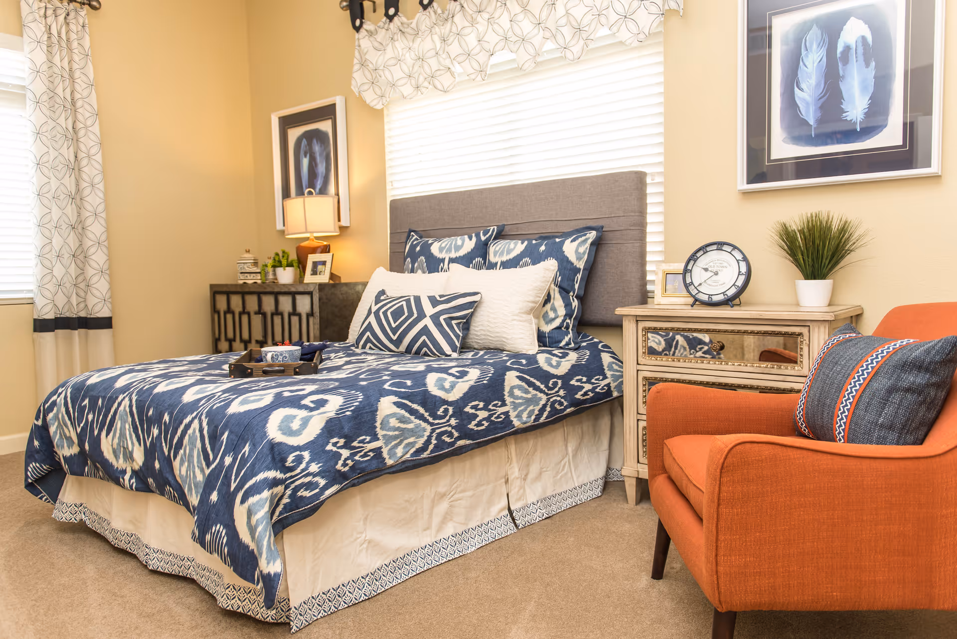 A cozy bedroom with a bed featuring blue and white patterned bedding and multiple pillows. Next to the bed is a wooden nightstand with a clock, a small plant, and a framed picture. An orange armchair with a decorative pillow is positioned beside the nightstand. The room has beige walls, a window with blinds and patterned curtains, and framed artwork on the walls.