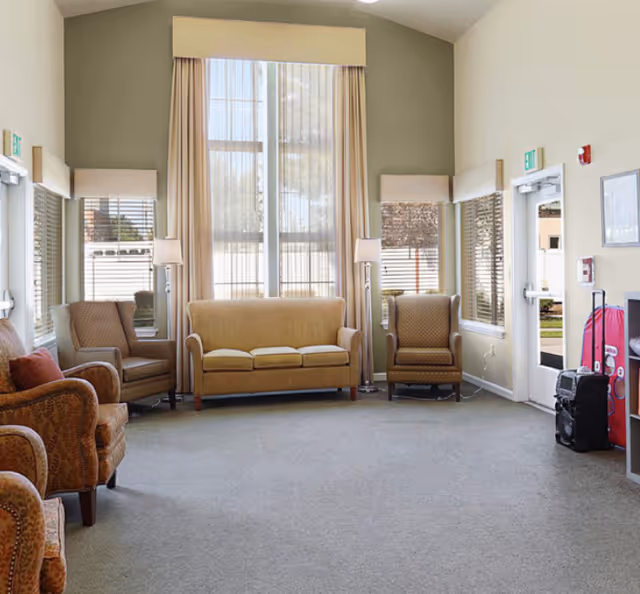A bright and spacious living room area with beige walls and carpet. The room features a beige three-seat sofa centered between two armchairs with patterned upholstery. There are two tall floor lamps on either side of the sofa. Large windows with sheer curtains allow natural light to fill the room. A door and additional windows are visible on the right side, along with some equipment and a red suitcase near the wall.
