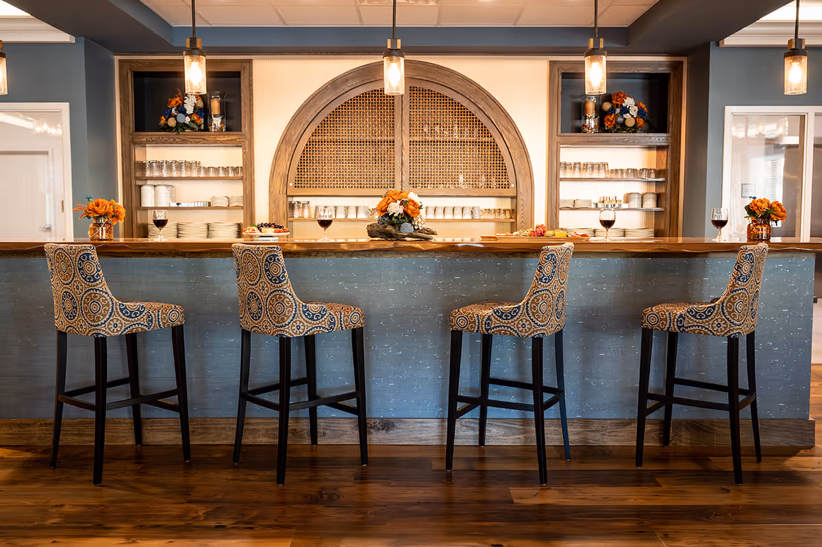A stylish bar area with four patterned high chairs lined up in front of a wooden counter. Behind the counter, shelves hold neatly stacked plates, glasses, and decorative flower arrangements. Three pendant lights hang from the ceiling, illuminating the space warmly.
