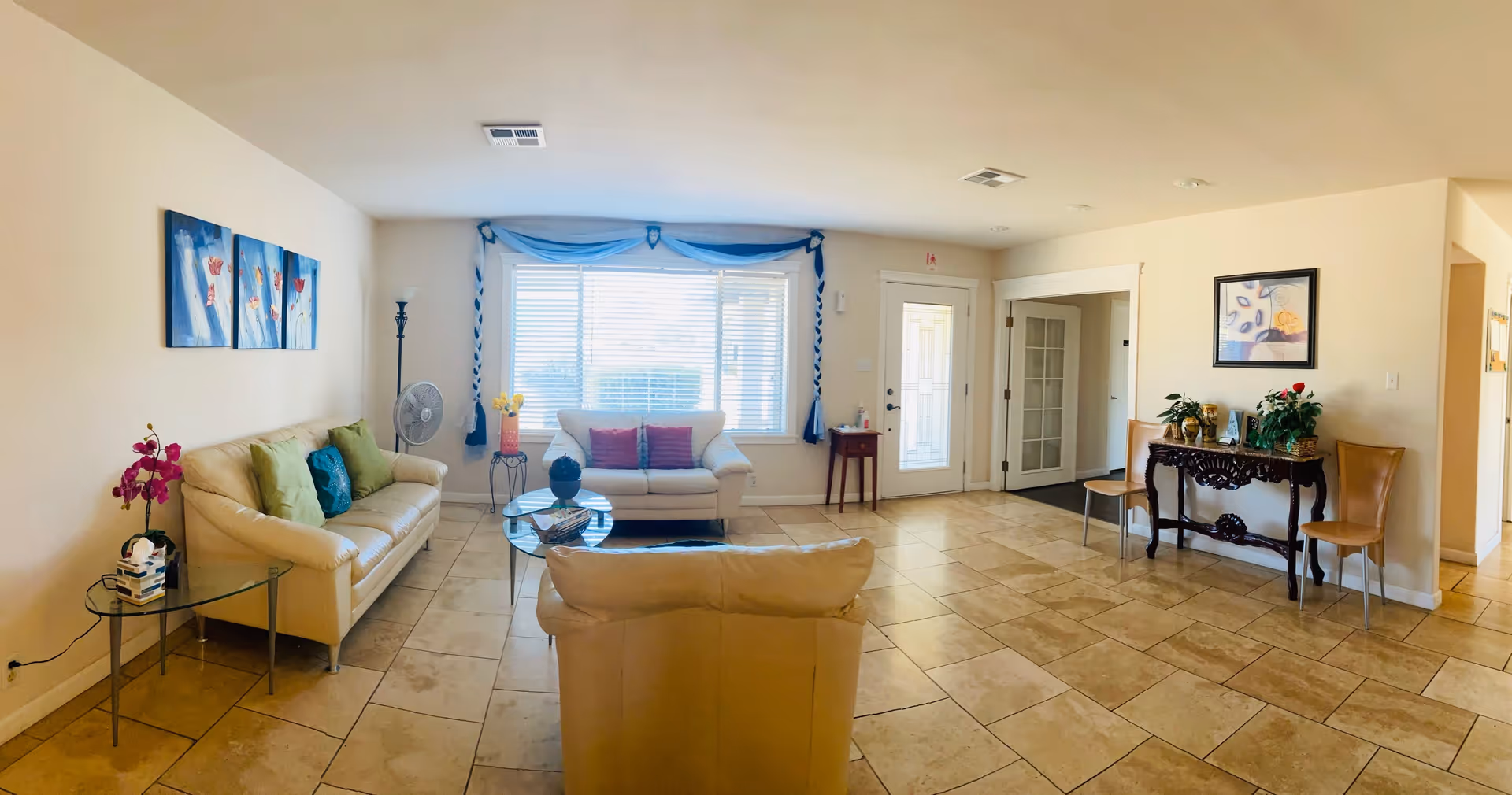 A bright and spacious living room with beige tiled floors and cream-colored walls. The room features a cream leather sofa with green and blue cushions, a matching loveseat with red cushions, and a single armchair. There is a glass coffee table in the center with decorative items, a side table with a pink orchid and a tissue box, and a tall floor lamp. A large window with blue curtains allows natural light to fill the room. On the right side, there is a decorative wooden console table with plants and framed artwork above it, flanked by two wooden chairs. The room has a door with glass panels and an adjacent hallway.