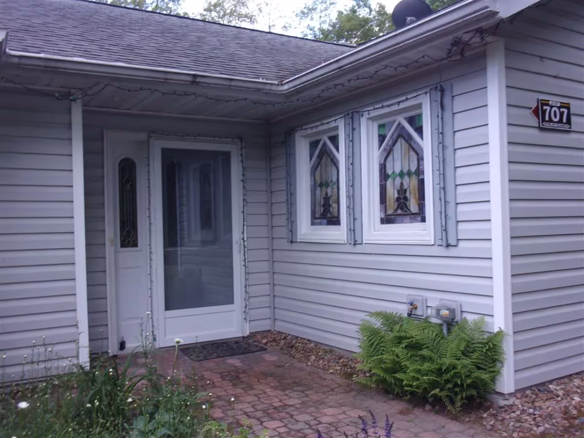 Front entrance of a light-gray siding building with a glass storm door, two decorative stained-glass windows, and a small brick walkway bordered by plants.
