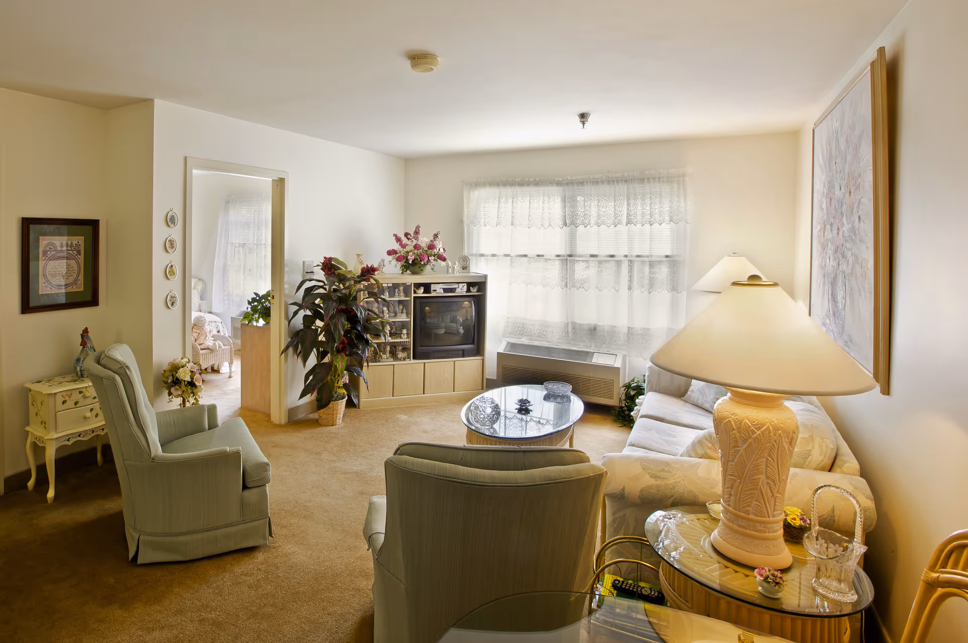 A cozy living room in Liberty Residence Independent Living featuring two upholstered armchairs, a floral-patterned sofa, a glass coffee table, a wooden entertainment center with a TV and decorative items, a large potted plant, and a table lamp on a glass side table. The room has beige carpeting, white walls, and a window with lace curtains letting in natural light.