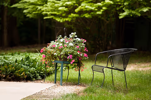A black metal outdoor bench next to a green metal plant stand holding a pot of pink and white flowers, set on a grassy area with trees and bushes in the background.