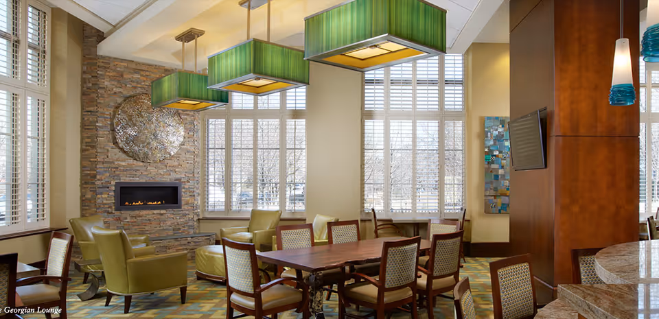 A cozy lounge area with a stone fireplace, green armchairs, and a wooden table surrounded by chairs. Large windows with white shutters let in natural light. Green rectangular pendant lights hang from the ceiling, and a flat-screen TV is mounted on a wooden column.