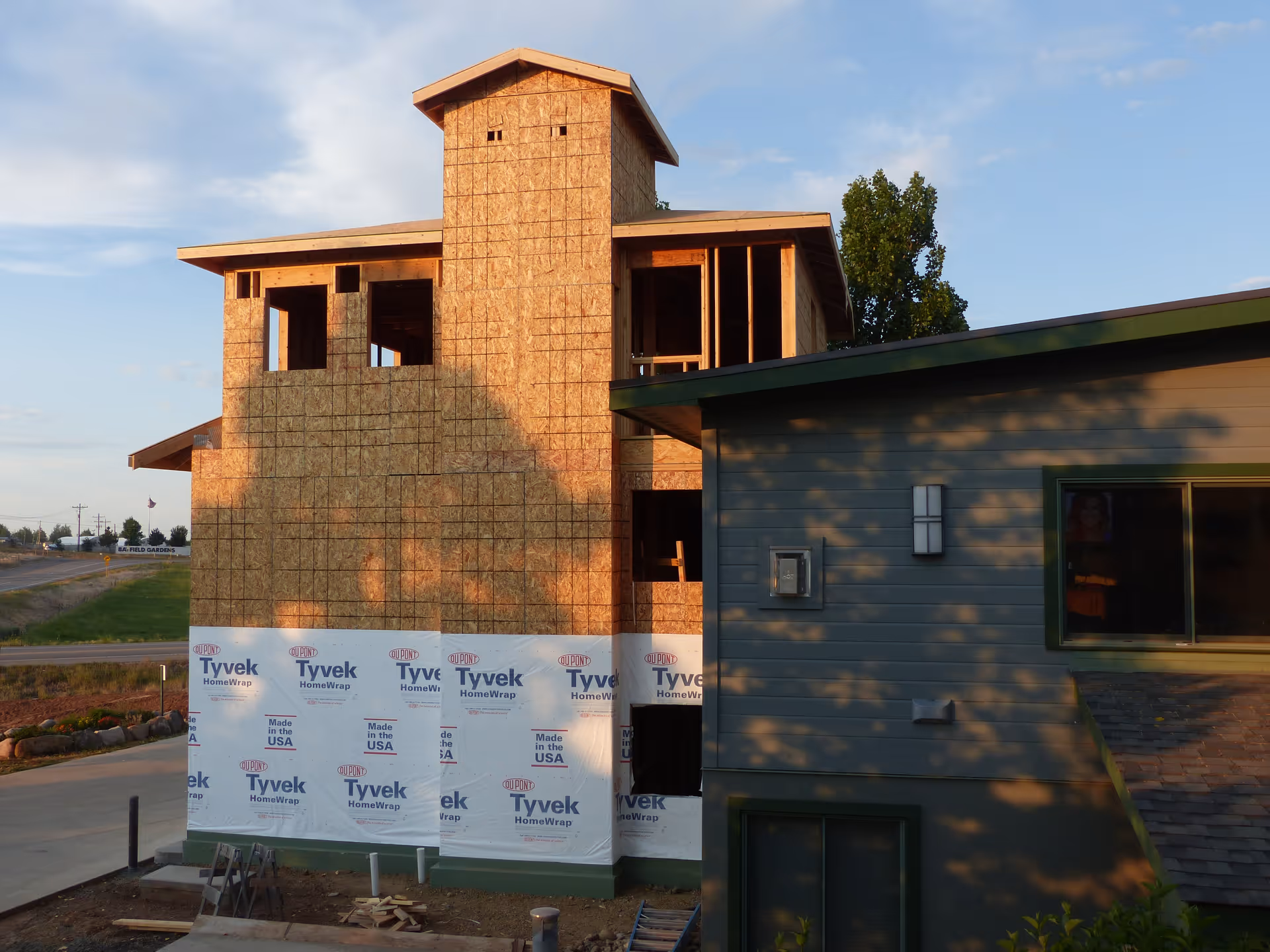 Partially constructed building with wooden framing and Tyvek HomeWrap on the lower section, next to a finished building with gray siding and green trim, under a partly cloudy sky.