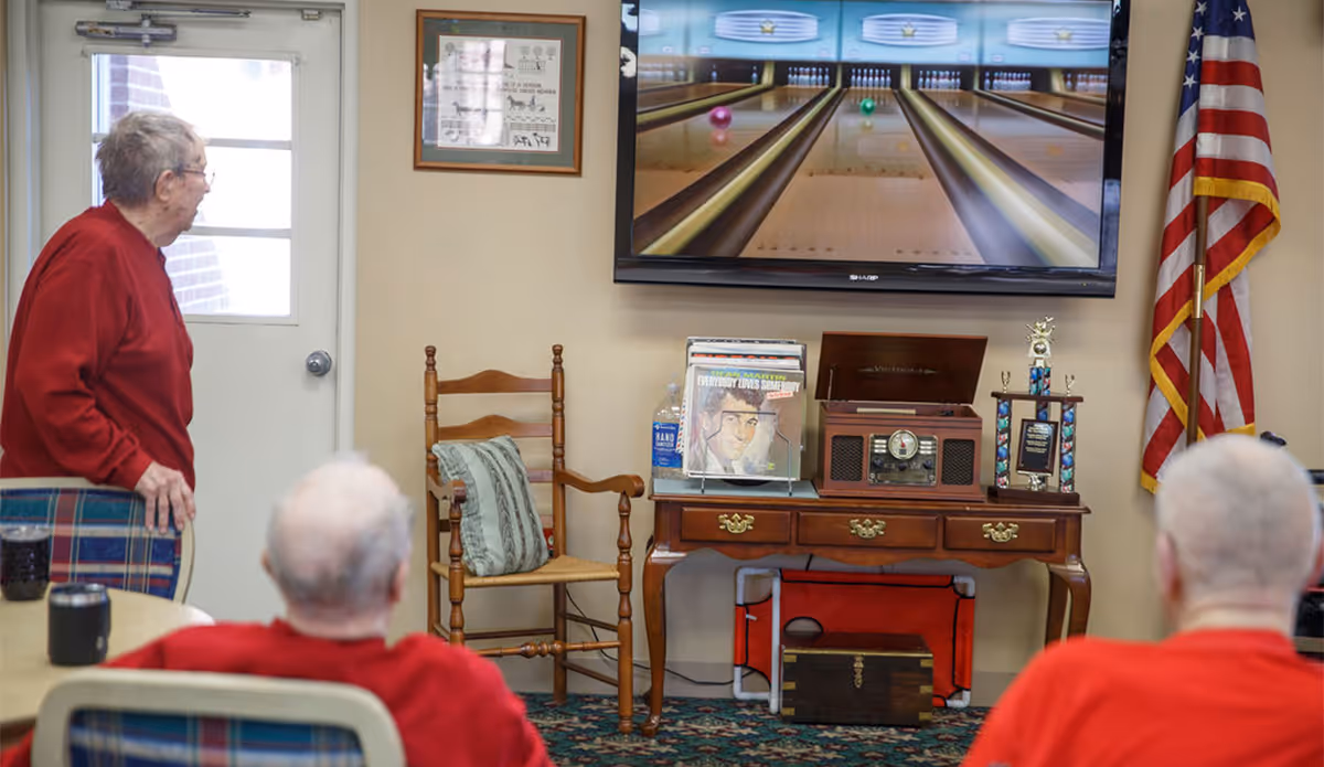 Three elderly people in a common room watching a bowling game on a large wall-mounted TV. The room has a wooden chair with a cushion, a wooden table with a vintage record player, records, a trophy, and an American flag in the corner.