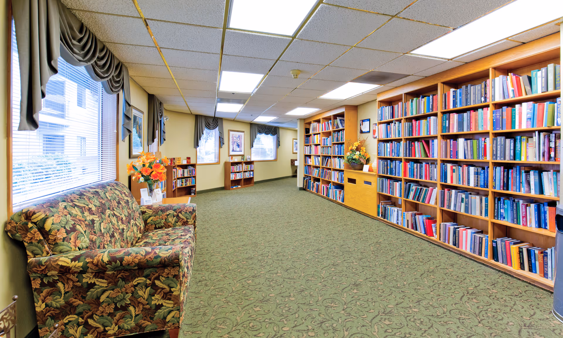 A cozy library room with large wooden bookshelves filled with books along the right wall. On the left side, there is a floral patterned couch beneath windows with draped curtains. The room has a green patterned carpet and bright fluorescent ceiling lights.
