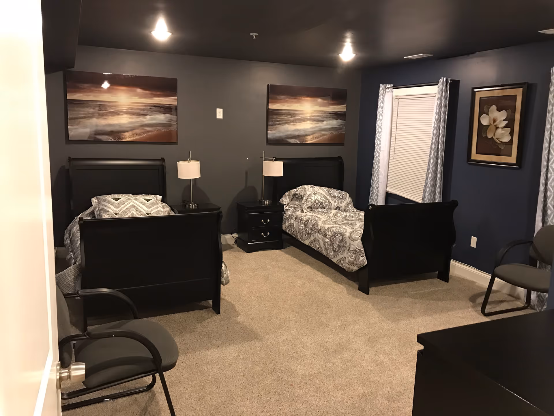 A bedroom with two single black wooden beds, each with patterned bedding. Between the beds is a black nightstand with a lamp. The walls are painted dark gray and blue, decorated with framed pictures of a sunset over the ocean and a flower. There are two gray chairs, one near the door and one near the window with closed blinds and patterned curtains. The floor is carpeted in beige.