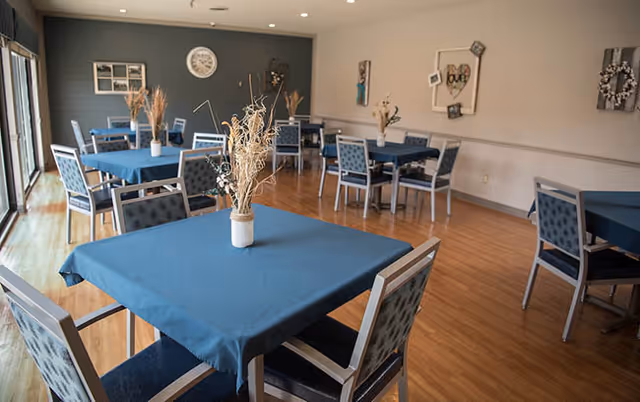 A dining room with multiple square tables covered with blue tablecloths, each surrounded by four chairs with patterned upholstery. The room has wooden flooring, light-colored walls, and large windows letting in natural light. Decorative vases with dried plants are placed on each table, and wall decorations including a clock and framed art are visible.