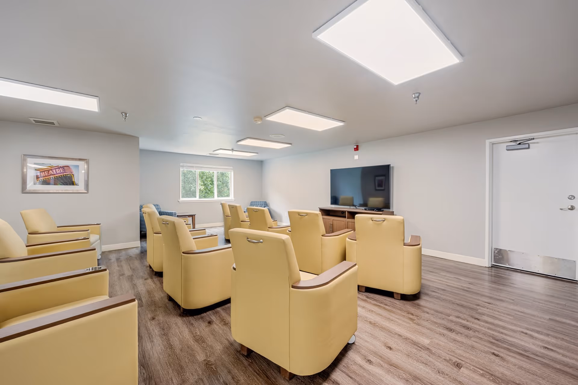 A common room with multiple beige armchairs arranged in rows facing a large flat-screen TV mounted on a wooden cabinet. The room has light gray walls, a window letting in natural light, wood flooring, and bright ceiling lights. There is a framed picture on the left wall and a white door on the right side.
