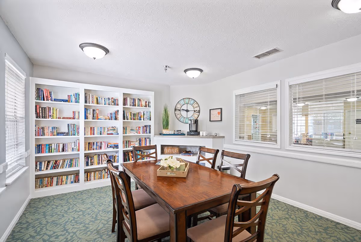 A bright room with a wooden table and six chairs, a large white bookshelf filled with books, two windows with blinds, a coffee maker on a small counter, and a decorative clock on the wall.