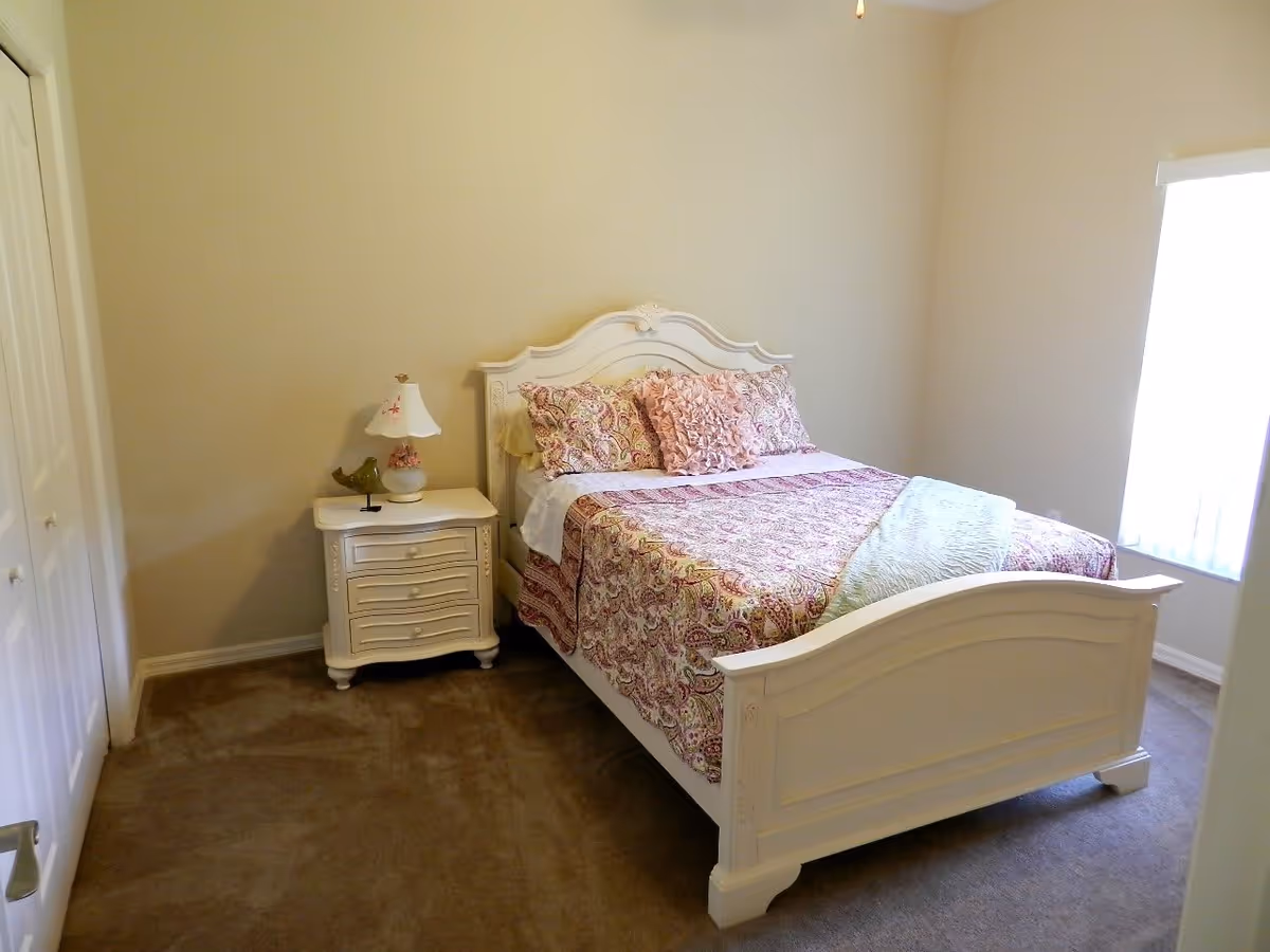 A bedroom with a white wooden bed frame and a bed made with a patterned pink and purple bedspread and matching pillows. There is a white nightstand next to the bed with a decorative lamp and a small bird figurine. The room has beige walls, a window with vertical blinds, and a carpeted floor.