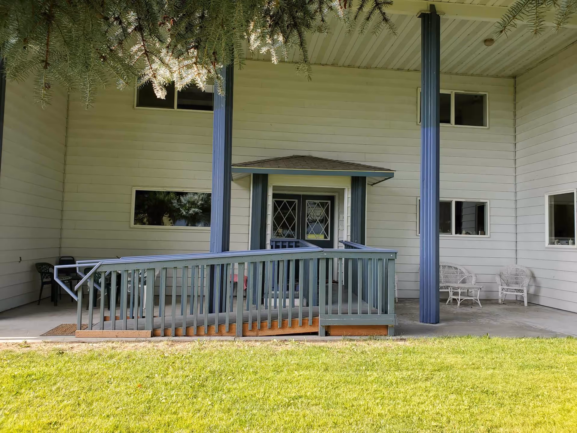 Covered front entrance of a light-colored building with a wheelchair ramp, blue support posts, and patio chairs.