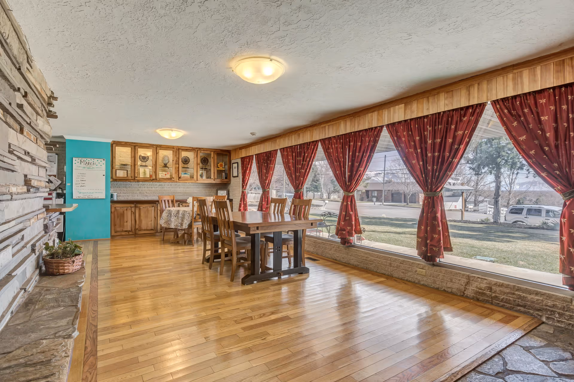 A spacious dining room with wooden floors and a large wooden dining table surrounded by chairs. The room features a stone fireplace on the left and large windows on the right with red curtains tied back, allowing natural light to fill the space. There are wooden cabinets and a small table with a floral tablecloth in the background.