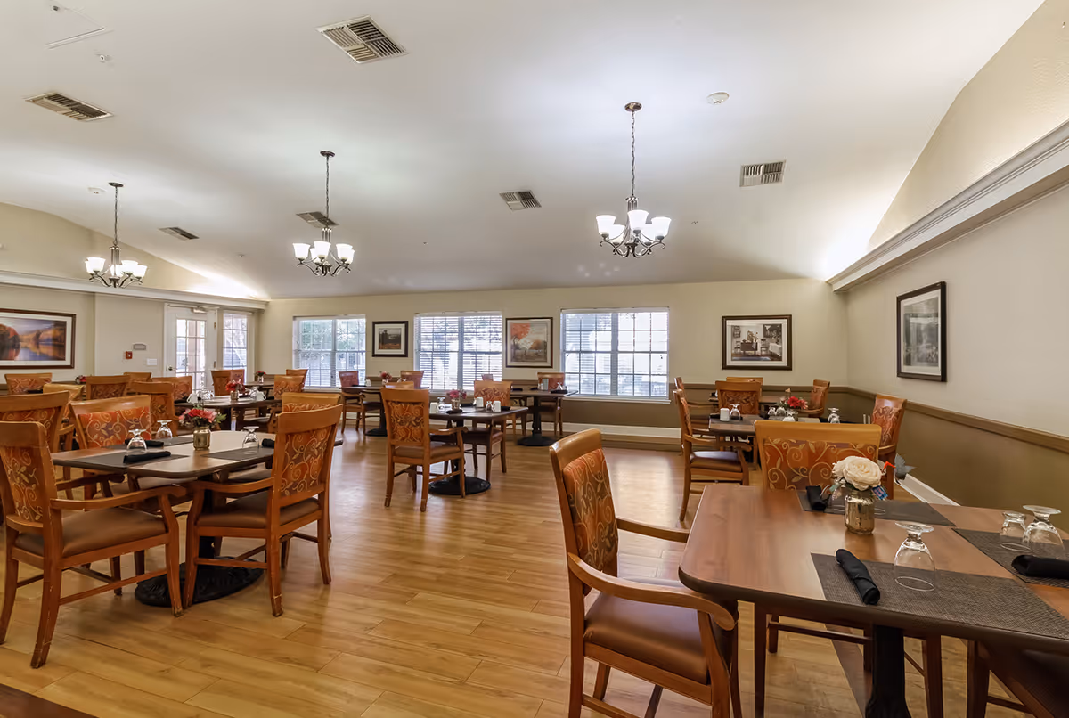 Spacious dining room with wooden tables and upholstered chairs arranged and set for meals, with chandeliers and large windows.
