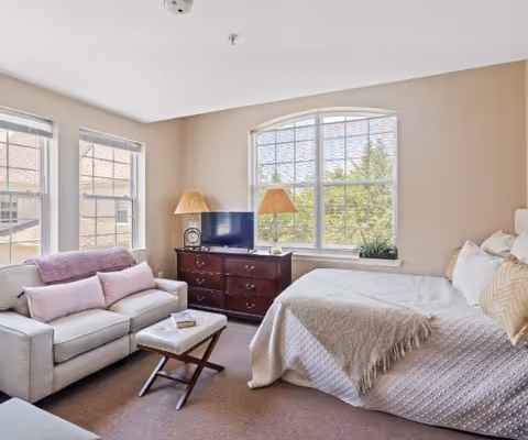A bright and cozy bedroom with a large window featuring white blinds. The room includes a bed with white bedding and multiple pillows, a wooden dresser with a small TV and two lamps on top, a beige sofa with pink cushions and a purple throw blanket, and a small cushioned ottoman in front of the sofa.