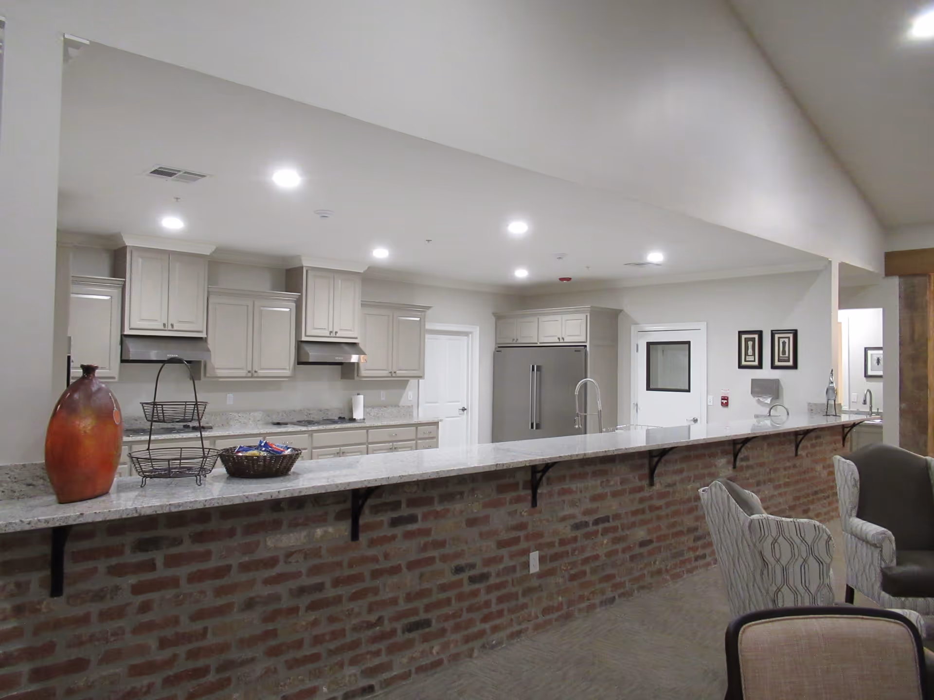 Interior view of a senior living facility kitchen with a long granite countertop supported by a brick base. The kitchen features light-colored cabinets, a stainless steel refrigerator, two range hoods, and a sink with a tall faucet. On the counter, there is a decorative vase, a two-tiered basket, and a basket with snacks. In the foreground, there are upholstered chairs and part of a dining or seating area visible.