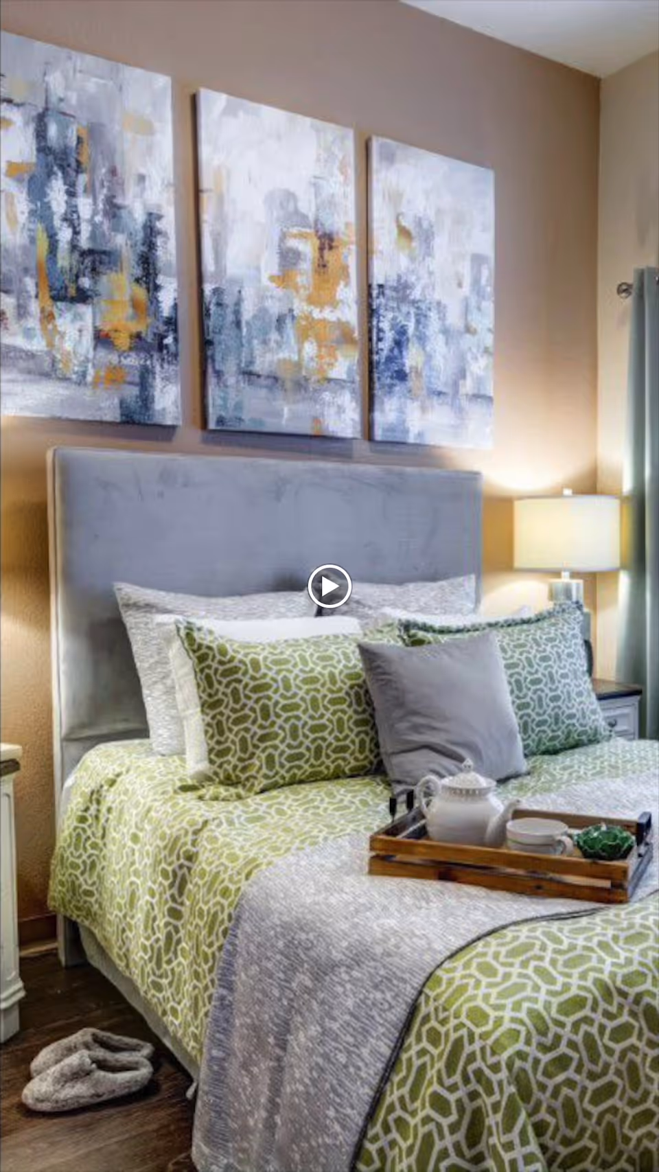 Staged bedroom with a gray upholstered headboard, green patterned bedding and pillows, a wooden tray with a teapot on the bed, a bedside lamp, and abstract triptych artwork above the headboard.