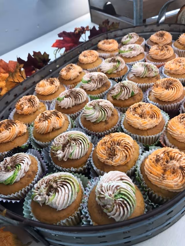 A tray filled with multiple cupcakes decorated with swirled frosting in shades of orange, white, and green, with some sprinkled with chocolate. The tray is placed on a white surface with autumn-themed decorations including artificial leaves in the background.