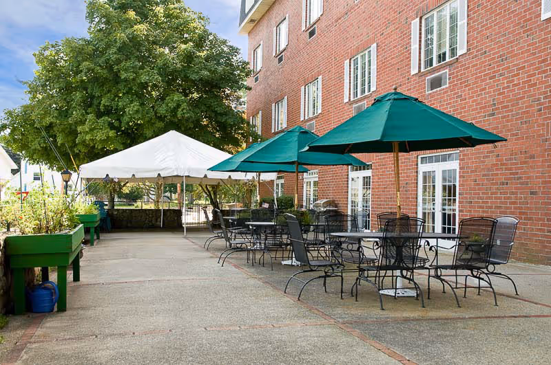 Outdoor patio area at a nursing and rehab center with several black metal tables and chairs, each table shaded by a green umbrella. There is a large white canopy tent in the background and a brick building with multiple windows and doors on the right side. Green planter boxes and a large tree are visible on the left side under a partly cloudy sky.