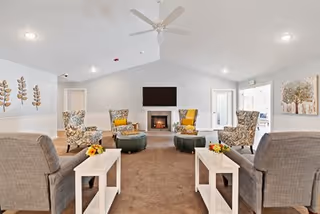 A bright and spacious living room with a vaulted ceiling, featuring a ceiling fan, a fireplace with a mounted flat-screen TV above it, and multiple upholstered armchairs arranged around two round ottomans. There are two side tables with flower arrangements, large windows letting in natural light, and decorative wall art on the walls.