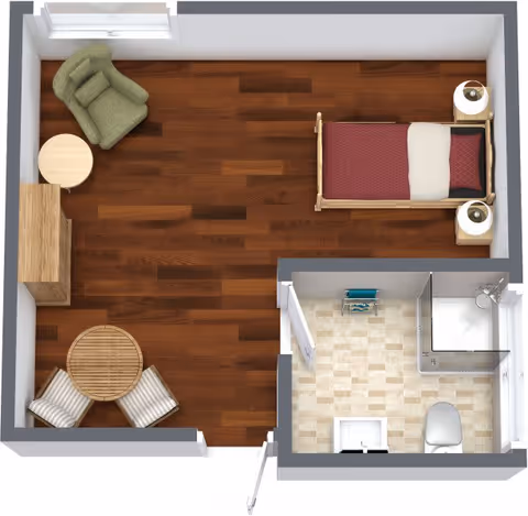 Top-down view of a small room layout with a single bed, two nightstands with lamps, a green armchair next to a round side table, a wooden cabinet, and a round dining table with two chairs. The room has wooden flooring. Attached is a small bathroom with tiled flooring, a shower, a toilet, and a sink.