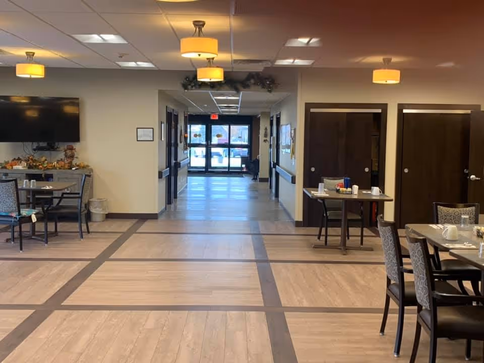 Interior view of a senior living facility dining area with tables and chairs arranged neatly. The floor has a light wood pattern with darker wood borders. There are ceiling lights with yellow lampshades and a hallway leading to glass exit doors in the background. A large flat-screen TV is mounted on the left wall above a counter with decorative items.