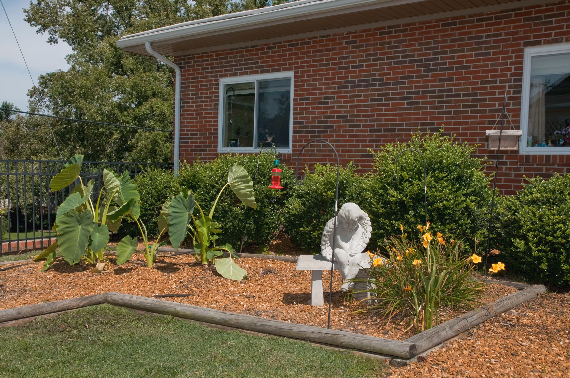 A garden area with a small statue of an angel sitting on a bench surrounded by green plants and yellow flowers. The garden is bordered by wooden logs and mulch, with a brick building and windows in the background.