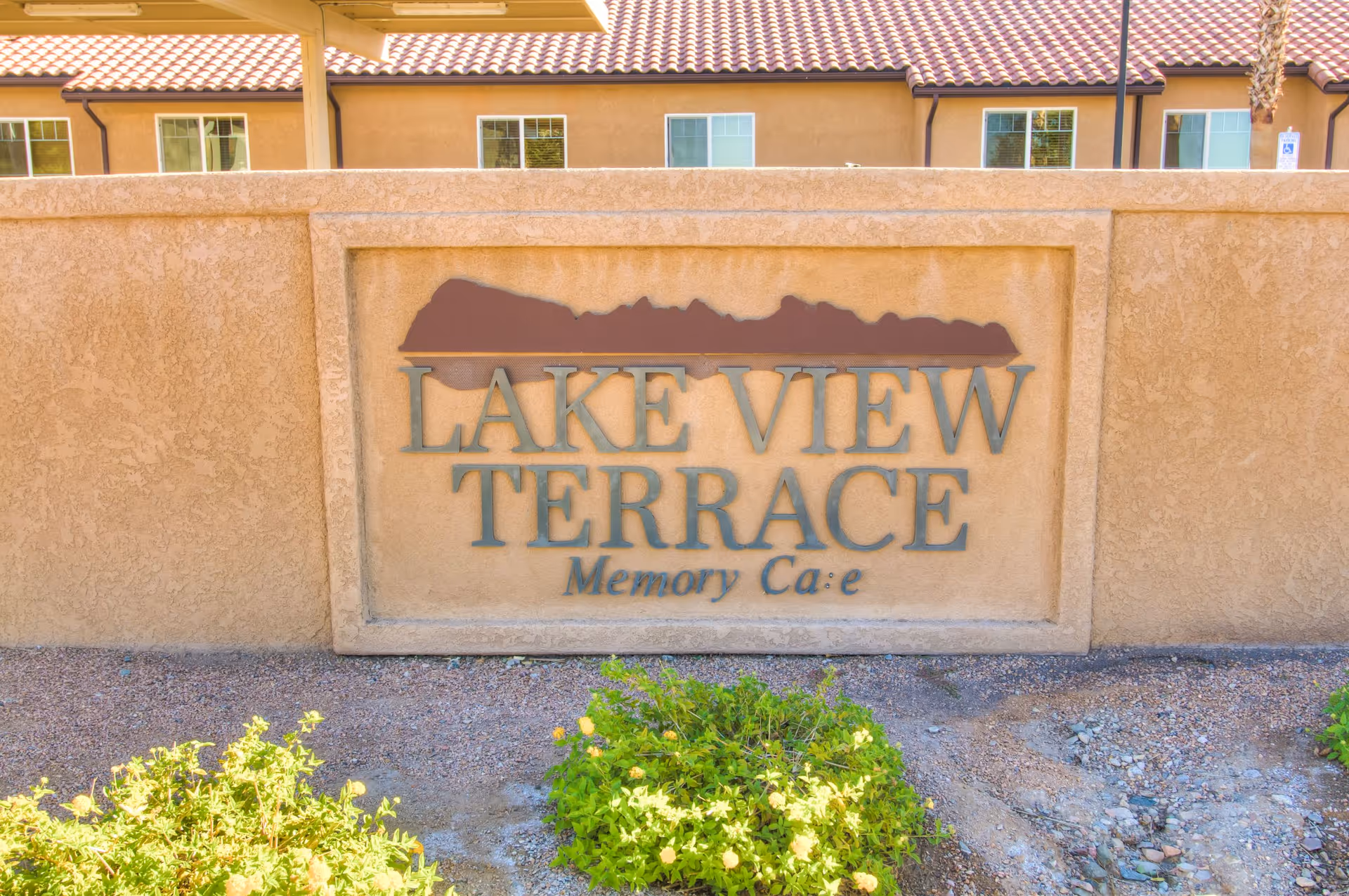 Entrance wall sign reading "LAKE VIEW TERRACE Memory Care" mounted on a stucco wall with the building and landscaping behind it.