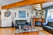 A cozy living room with two black leather sofas, a wooden coffee table with decorative items, a fireplace with a wooden mantle, wall art including a large painting and decorative plates, and a large window with green curtains letting in natural light.