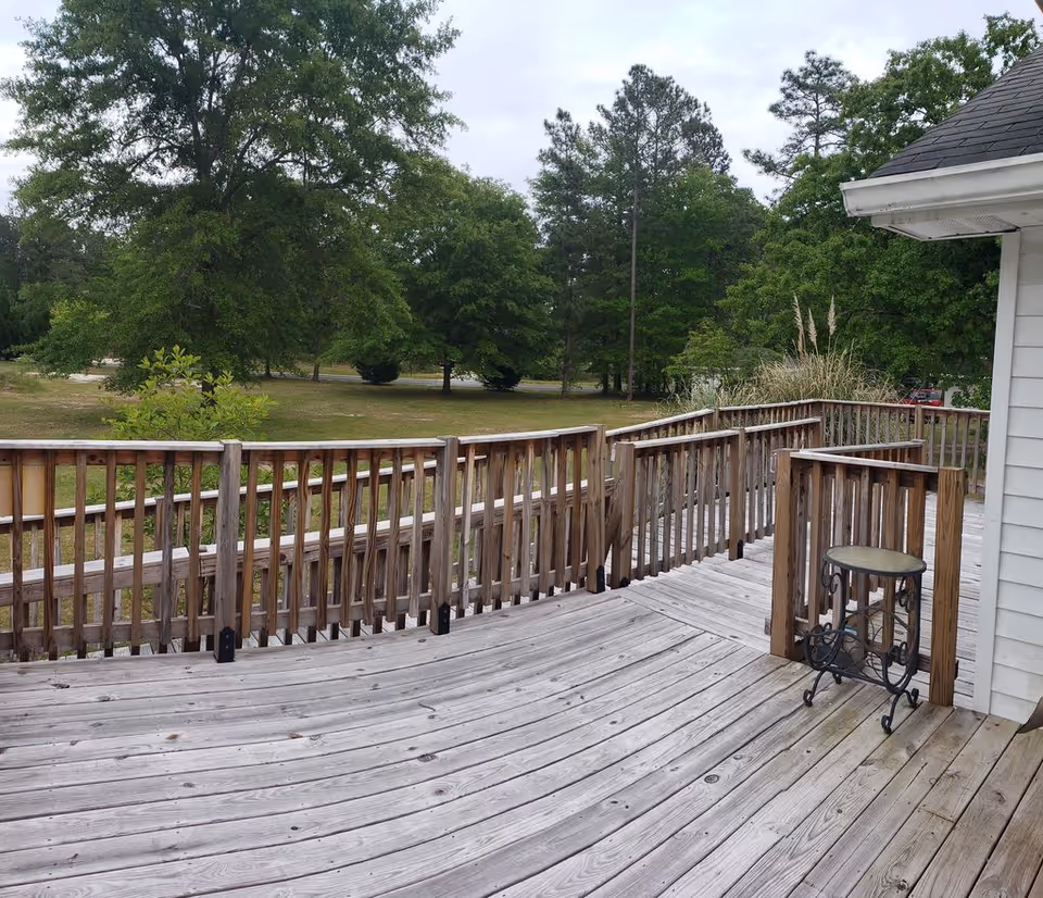 Wooden outdoor deck with railings and a small round glass-top table, overlooking a grassy area with trees under a cloudy sky.