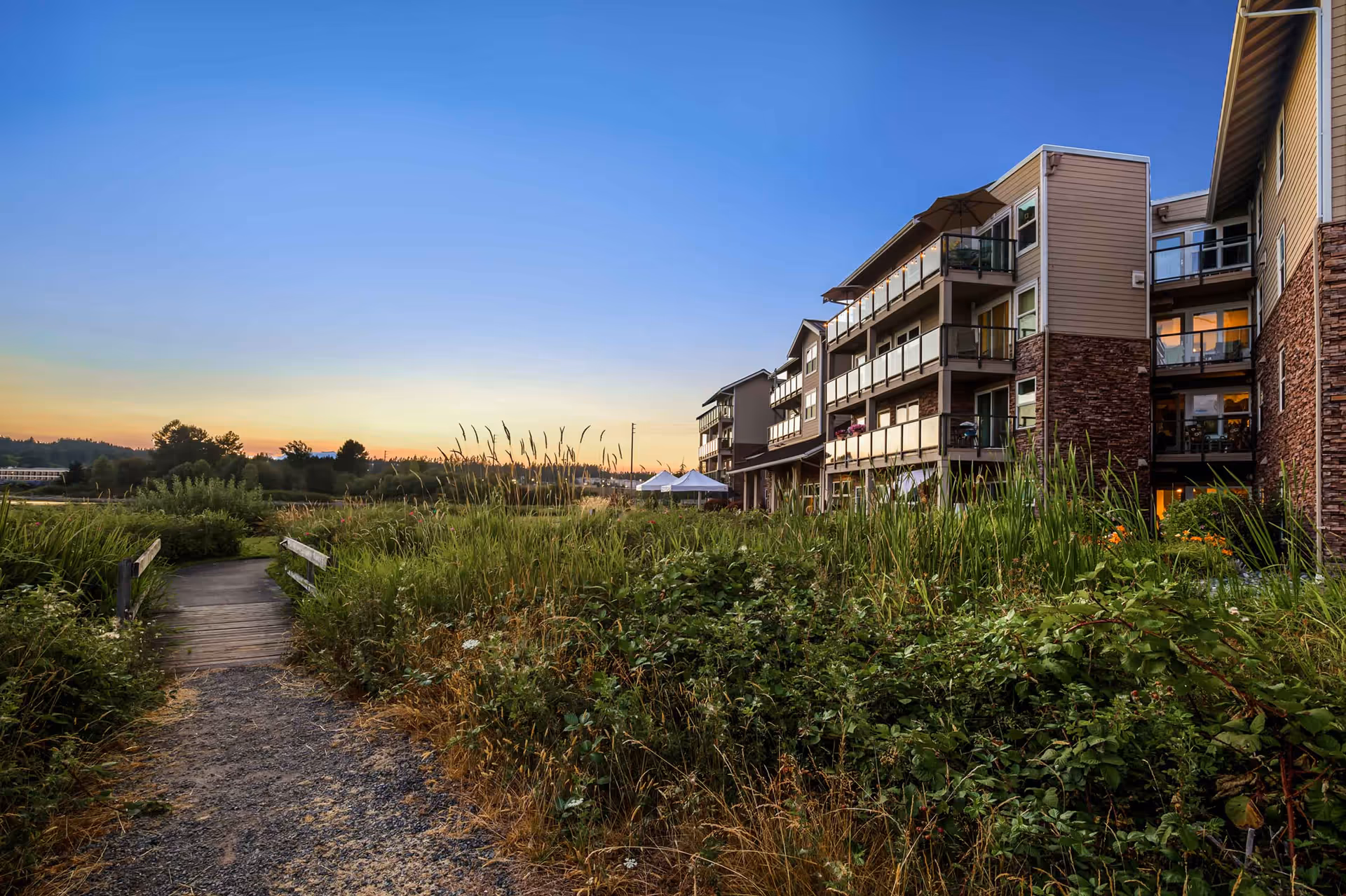A gravel path and small boardwalk through tall grasses leading to a multi-story senior living building at sunset.