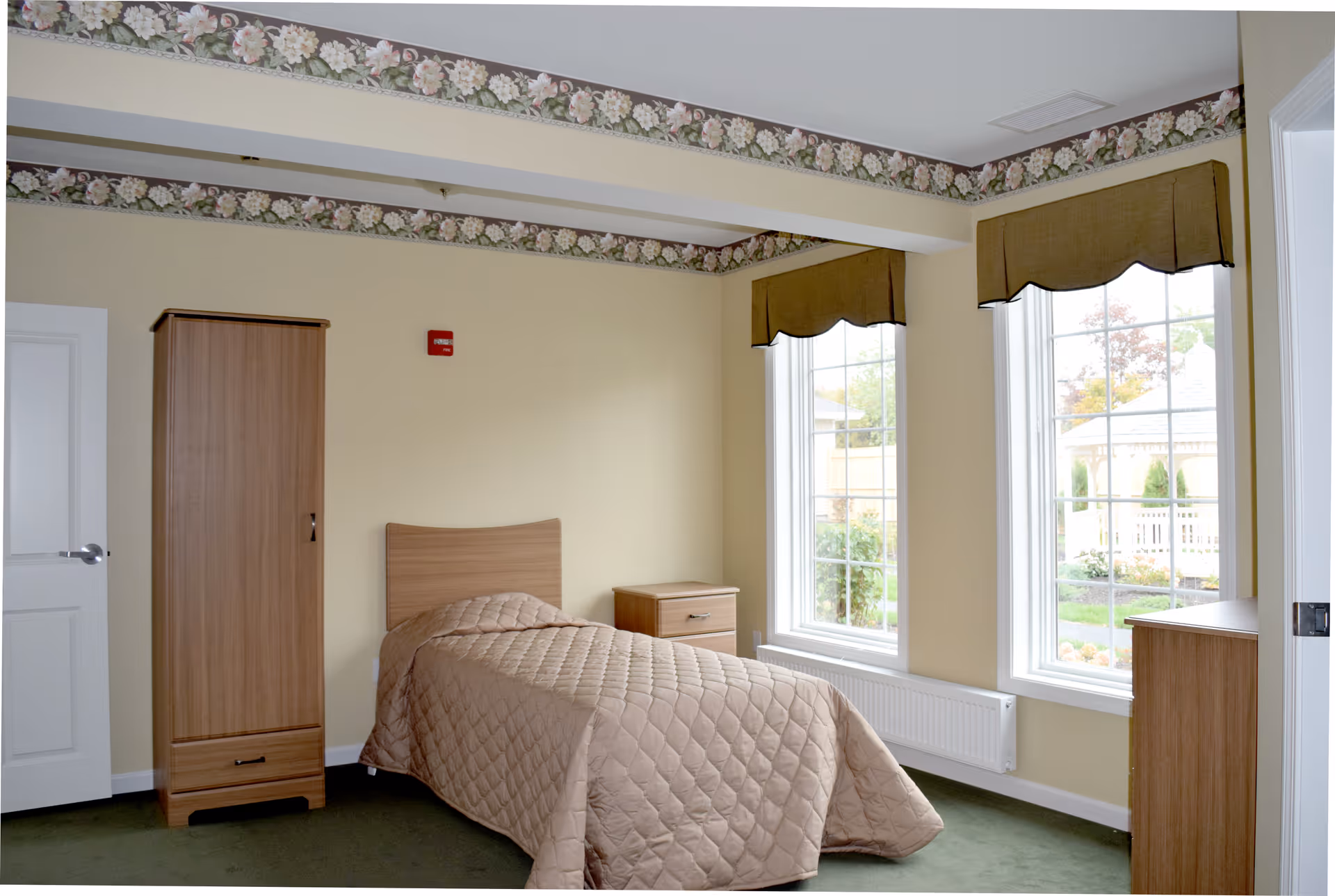 A simple single bedroom with a bed, wooden wardrobe and dresser, and two tall windows letting in natural light.