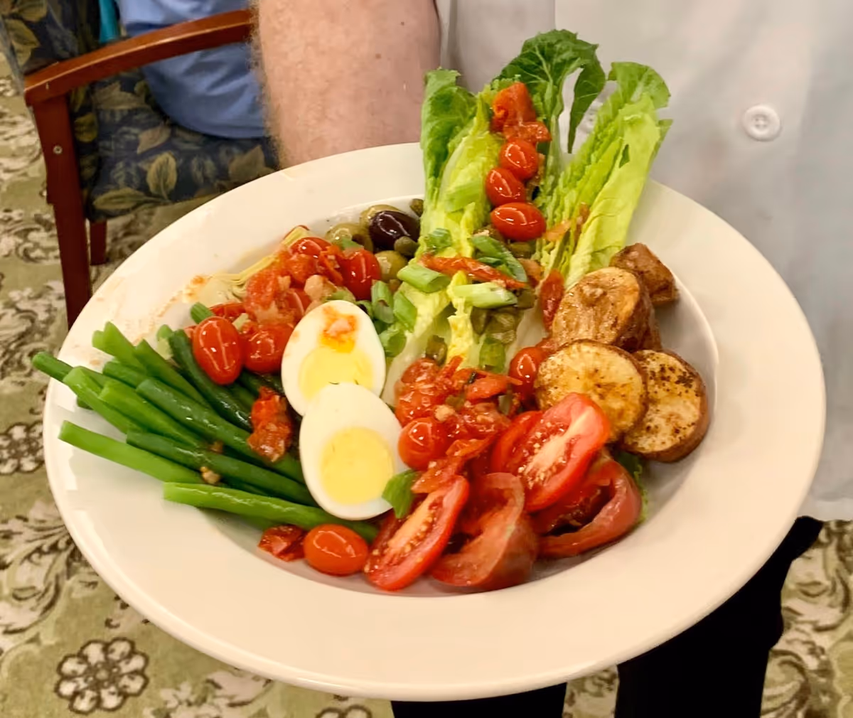 A white plate filled with a variety of fresh foods including green beans, halved hard-boiled eggs, cherry tomatoes, sliced tomatoes, lettuce leaves topped with chopped vegetables, and roasted potato slices. The plate is being held by a person wearing a white shirt, with a patterned carpet and a chair visible in the background.