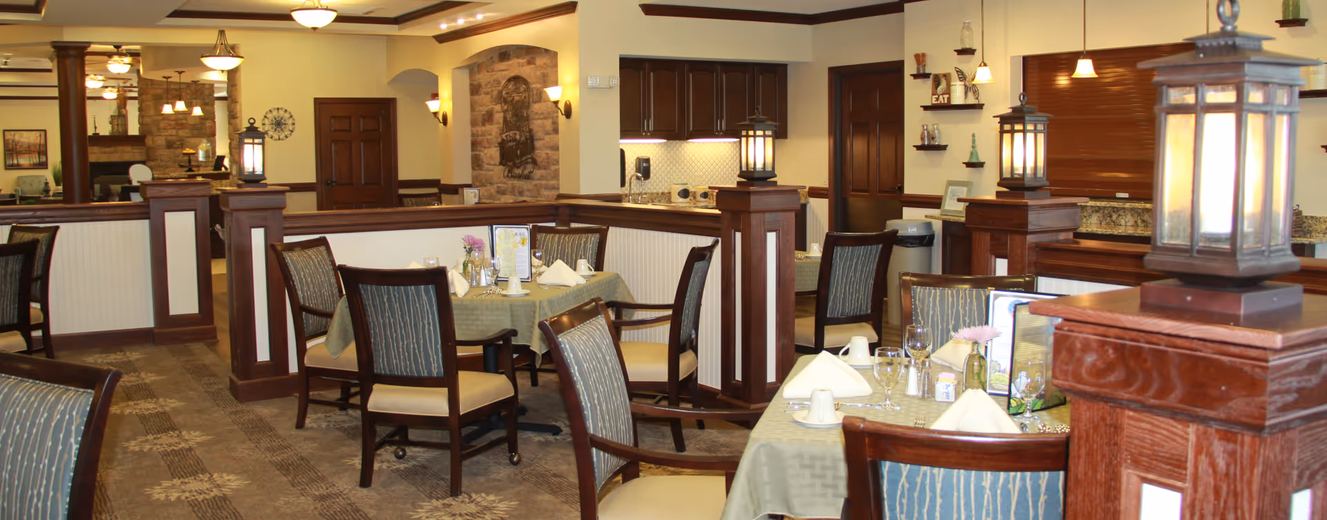 Communal dining room with neatly set tables, upholstered chairs, and decorative wooden posts and lamps.