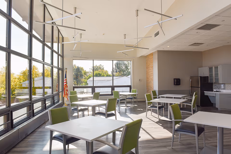 A bright dining room with large floor-to-ceiling windows letting in natural light. The room contains several square white tables with green and gray chairs arranged around them. There is an American flag in the corner near the windows, and a kitchenette area with a refrigerator, sink, and cabinets along one wall.