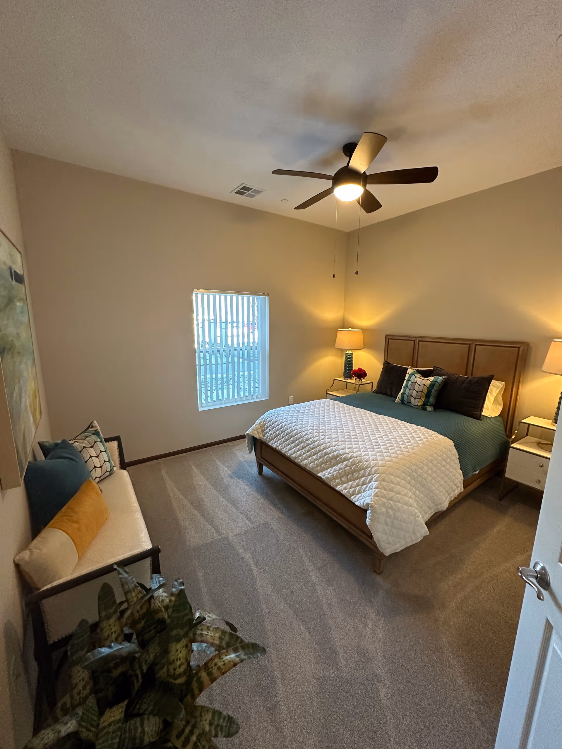 A cozy bedroom with a large bed covered in a white quilt and teal bedding, two nightstands with lamps on either side, a ceiling fan with light, a small bench with colorful pillows, a window with vertical blinds, a plant in the foreground, and neutral-colored walls and carpet.