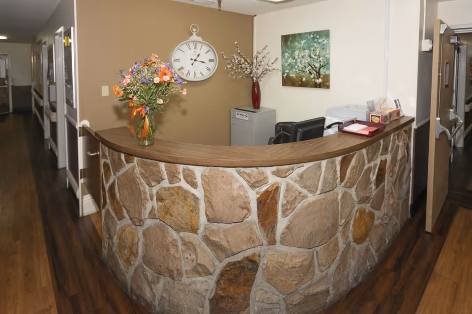 Reception desk area with a stone facade and wooden countertop in a healthcare facility. On the desk, there is a vase with colorful flowers and some office supplies. Behind the desk, a wall clock and a painting of a flowering tree are visible. The floor is wooden, and hallways extend on both sides of the desk.