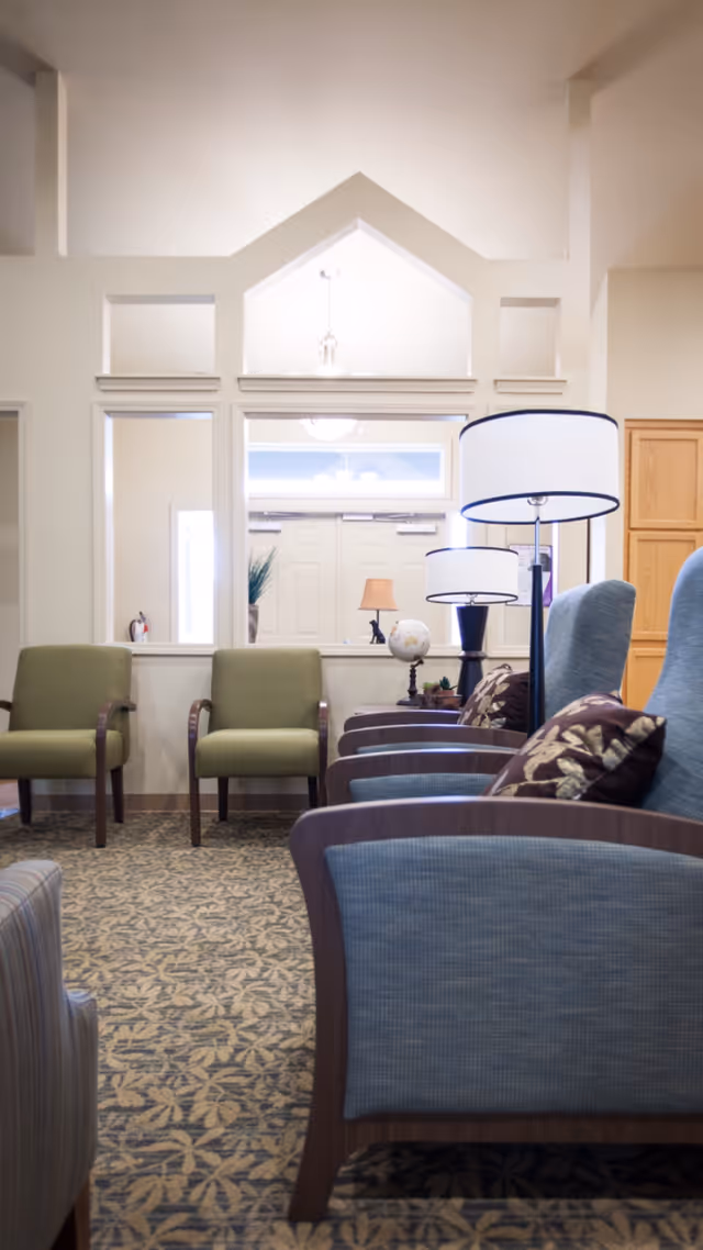 A cozy seating area in a memory care facility with several upholstered chairs arranged in rows. The chairs have wooden armrests and some have patterned cushions. There are two floor lamps with white shades providing soft lighting. The room has a patterned carpet and a large window with a view into another room with a door and additional lamps.