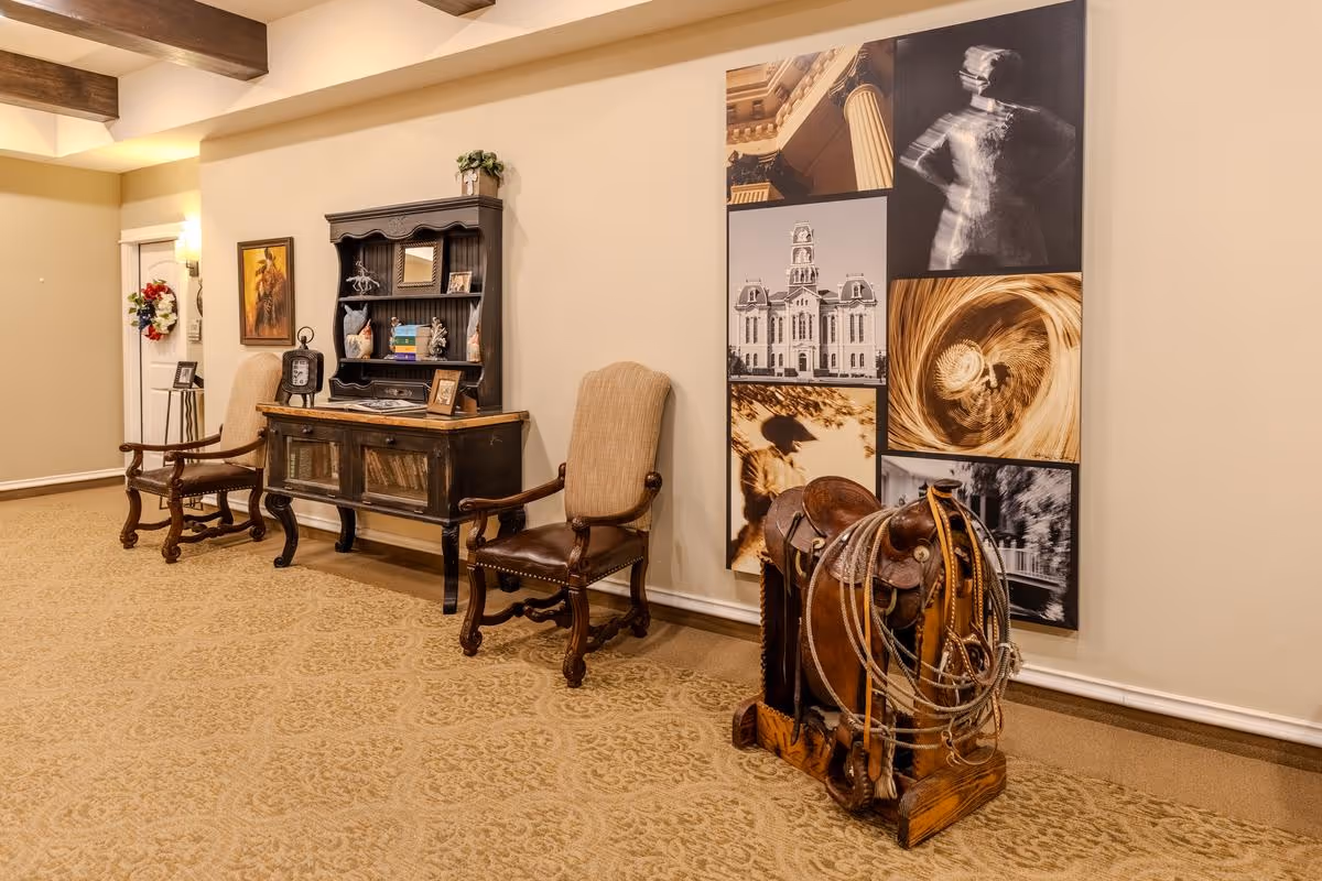 A hallway in an assisted living facility featuring two wooden armchairs with beige cushions, a dark wooden cabinet with decorative items and books, a wall adorned with a collage of black and white and sepia-toned photographs, and a wooden saddle with ropes displayed on the floor.