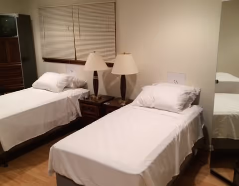 A bedroom with two single beds covered in white linens, each with a pillow. Between the beds is a wooden nightstand with two matching table lamps. A window with closed blinds is above the nightstand. A mirror is visible on the right side of the image reflecting part of one bed.