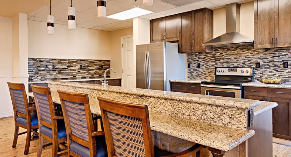 Modern kitchen with a granite island, wooden barstools, stainless steel appliances, and a mosaic tile backsplash.
