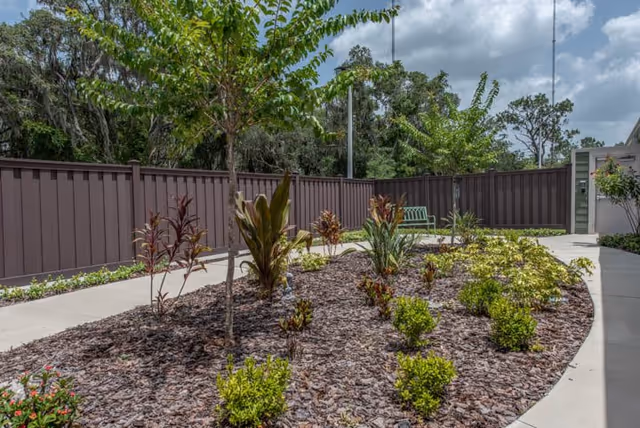 Outdoor garden area with a variety of small plants and shrubs planted in mulch beds, surrounded by a concrete walkway. There is a brown fence enclosing the area and a green bench near the back. Trees and a partly cloudy sky are visible in the background.